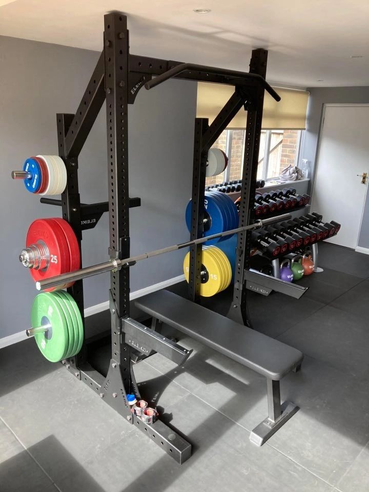Squat rack with a loaded barbell and red, blue, green and yellow bumper plates racked either side, dumbbell rack behind, black rubber floor.