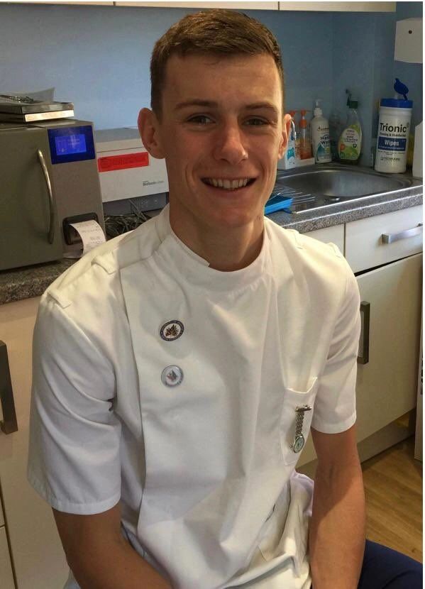 Jamie Bedwell in his clinic room in a white tunic, smiling, autoclave and worktop behind him.