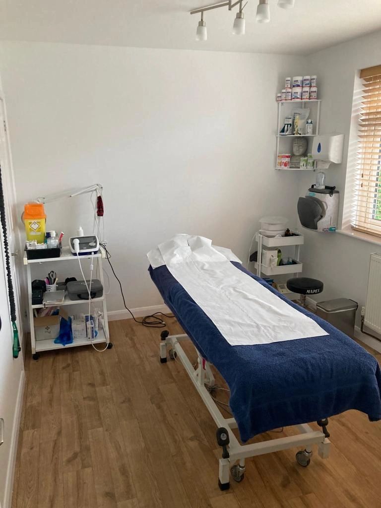 Karen's treatment room at 2 Church Road: a navy-covered couch with a white paper roll, pale oak floor, electrotherapy trolley and a venetian blind.